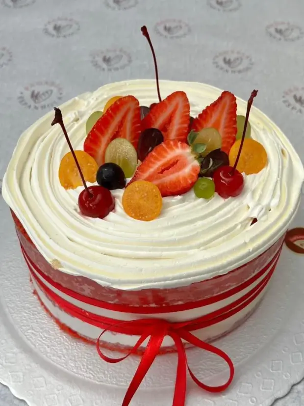 Bolo de chantilly com morangos, uvas e cerejas por cima e fita vermelha na lateral
